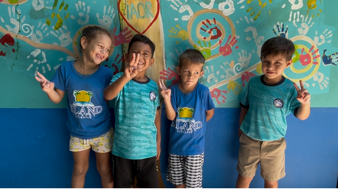 Four young IAOR students posing in front of the school's colorful handprint mural